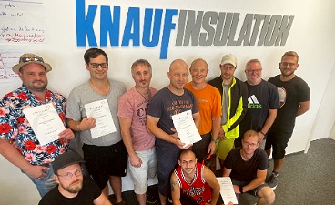 Gruppenbild nach einer Mitarbeiterschulung vor dem Firmenschriftzug Knauf Insulation. Mehrere Personen halten Teilnahmeurkunden in der Hand.