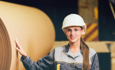 Frau mit Helm steht in Fabrick vor Papierrolle 